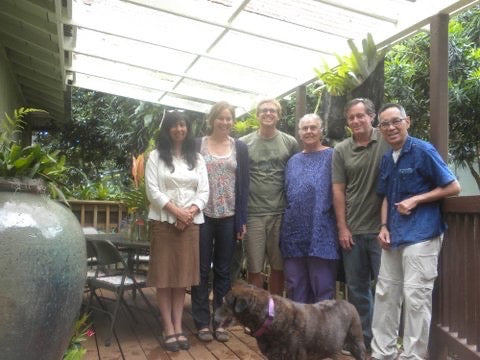 Group of people standing outdoors on a wooden deck with plants and a dog.