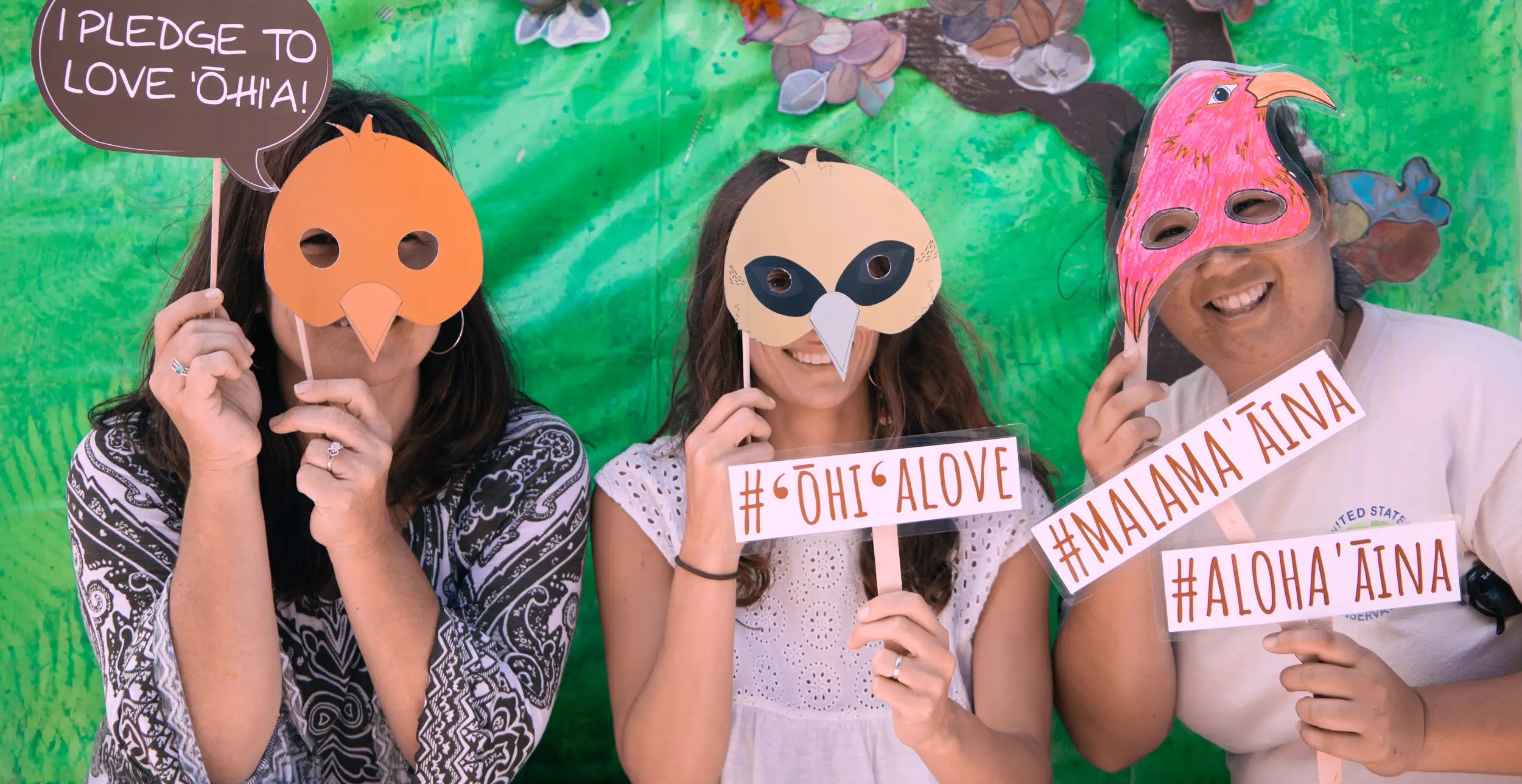 Three people wearing colorful masks and holding signs with Hawaiian phrases against a green background.